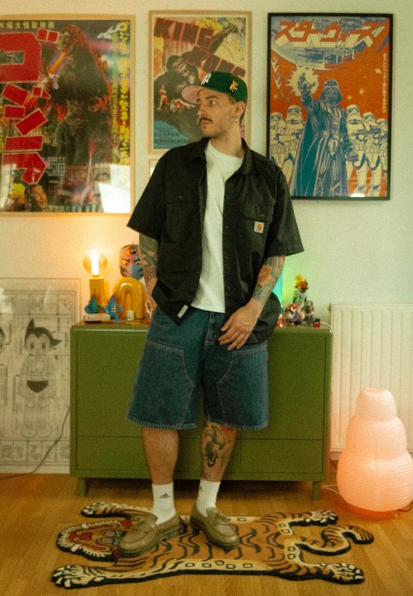 Tatuoitu mies, joka käyttää lippalakkia, mustaa paitaa, valkoista t-paitaa ja farkkushortseja, seisoo tiikerin muotoisella matolla huoneessa, jossa on kehystettyjä julisteita ja vihreä kaappi.