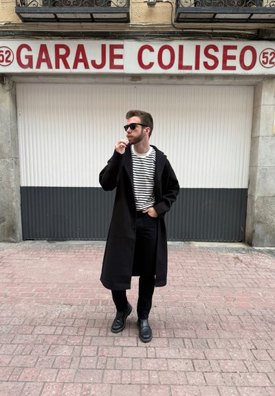 Largo abrigo negro sobre una camisa de rayas blancas y negras, combinado con pantalones y botas negras. El fondo presenta una puerta de garaje blanca con texto rojo.