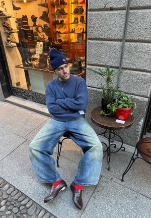 Hombre con gorro azul, suéter, pantalones anchos, calcetines rojos y mocasines sentado junto a un muro de piedra y una mesa con macetas fuera de una tienda de zapatos.