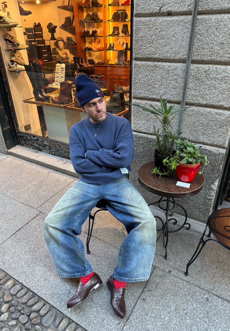 Homme portant un bonnet bleu, un pull, un jean large, des chaussettes rouges et des mocassins, assis à côté d’un mur en pierre et d’une table avec des plantes en pot devant un magasin de chaussures.