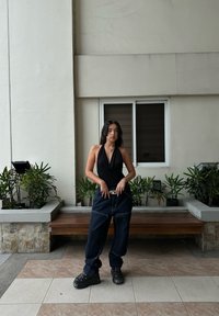 Schwarzes Halftertop, lockere dunkelblaue Jeans, schwarze Plateausneaker. Die Szene zeigt eine strukturierte Wand und Topfpflanzen im Hintergrund.
