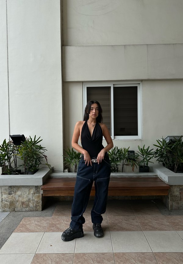Sort haltertop, løse mørkeblå jeans, sorte platform sneakers. Scenen har en tekstureret væg og potteplanter i baggrunden.