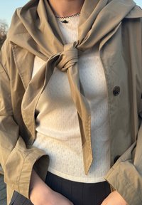 Μπεζ ελαφρύ μπουφάν δεμένο στο λαιμό πάνω από ένα λευκό υφαντό μπλουζάκι με μικρά σχέδια και μαύρο κουμπί. Είναι ορατό ένα σκούρο πιτζάμι με πτυχώσεις.