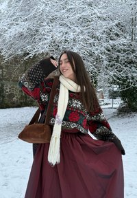 Ung kvinde i mønstret sweater og hvidt tørklæde poserer smilende i snedækket landskab med bare, snebeklædte trægrene bagved.