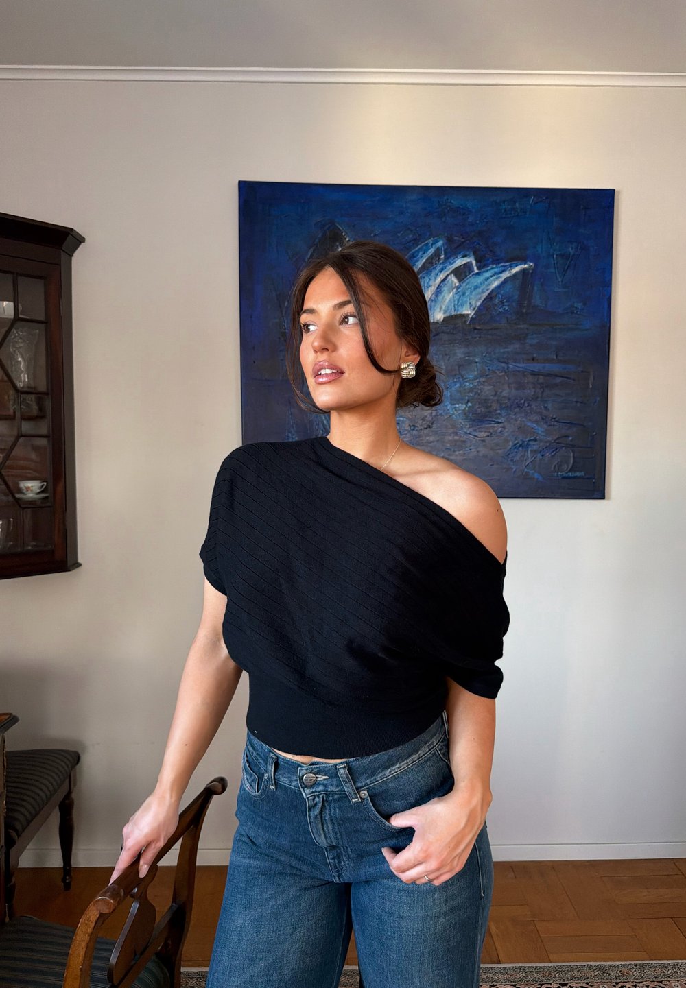 Jeune femme aux cheveux bruns attachés en chignon, portant un haut noir décolleté et un jean bleu, debout à l'intérieur près d'une chaise en bois et d'une œuvre murale bleue.