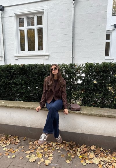 Chaqueta marrón de doble botón sobre una blusa oscura, vaqueros acampanados azules, bolso marrón, zapatillas grises, sentado en un banco de piedra con hojas de otoño alrededor.