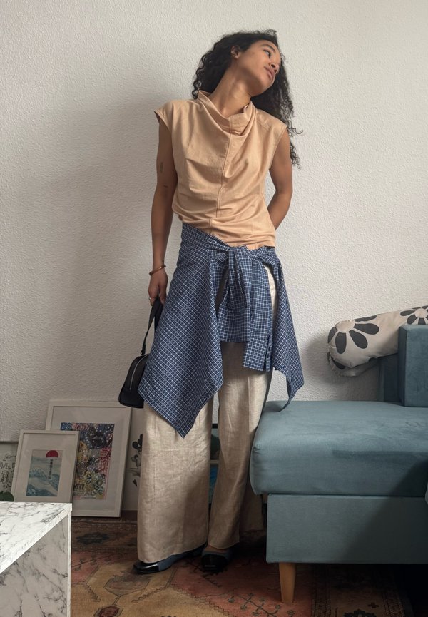 Femme aux cheveux bouclés, debout de manière décontractée contre un mur blanc, portant un haut beige sans manches, un pantalon large, avec une chemise à carreaux bleue nouée autour de la taille.