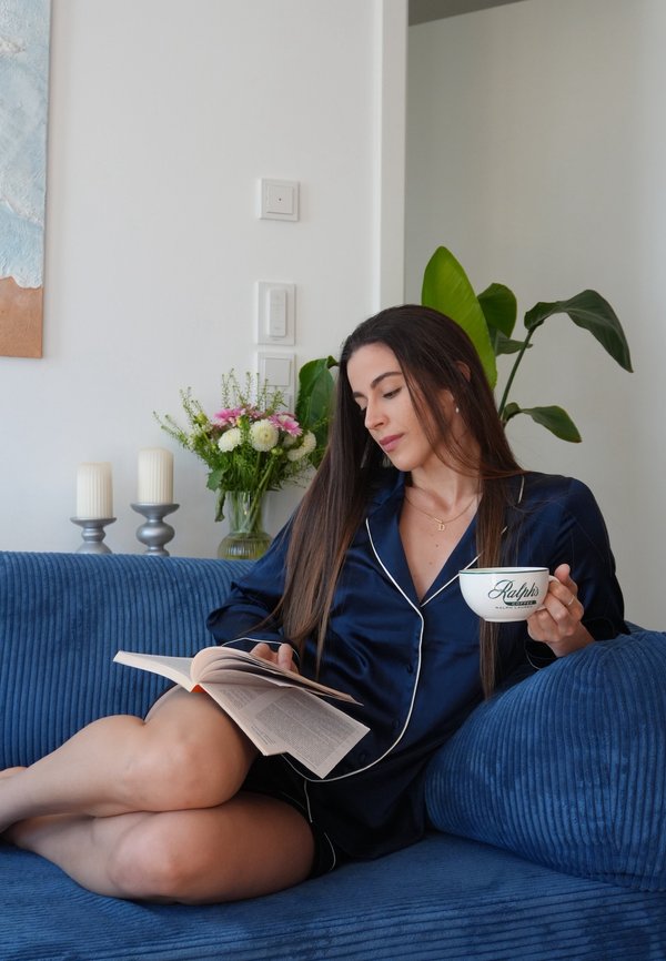 Mujer con pijama azul marino sentada en un sofá azul, leyendo un libro y sosteniendo una taza blanca de café con una planta y velas de fondo.