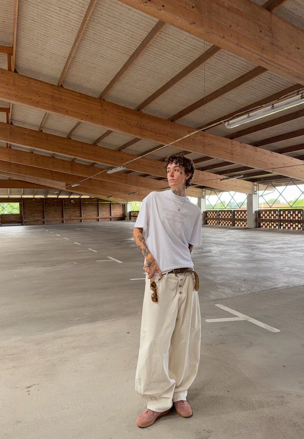 Person mit Tattoos, die übergroße weiße Hosen und ein weißes T-Shirt trägt, steht in einer leeren, überdachten Tiefgarage mit Holzbalken.