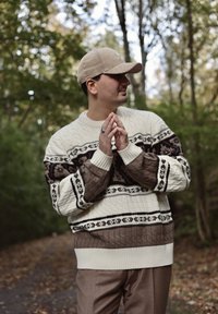 Man in a beige cap, patterned cable-knit sweater, and brown trousers stands on a forest path with his hands clasped, looking to the right.