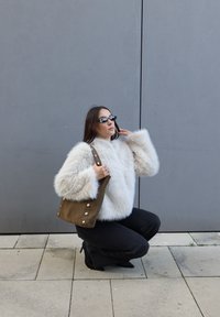 Femme portant un manteau en fausse fourrure blanche, un pantalon noir et des lunettes de soleil, accroupie et tenant un sac à main marron devant un mur gris.