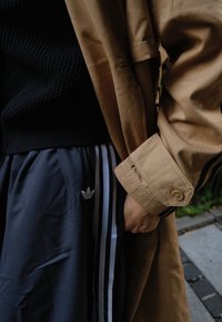 Manteau trench-coat beige porté sur un pull en maille noir et un pantalon de sport bleu marine avec des rayures blanches et un logo Adidas. Texture lisse et détails de boutons visibles.