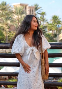 White eyelet dress with puff sleeves and a tied neckline, featuring intricate cutouts; accessorised with a woven bag and delicate jewellery.