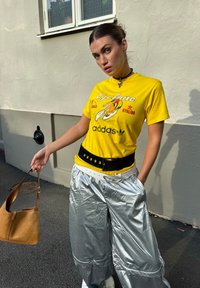 Žlté bavlnené tričko s grafikou cyklistiky a logom „Adidas“, doplnené lesklými striebornými oversized nohavicami a čiernym opaskom. Hnedá kabelka.