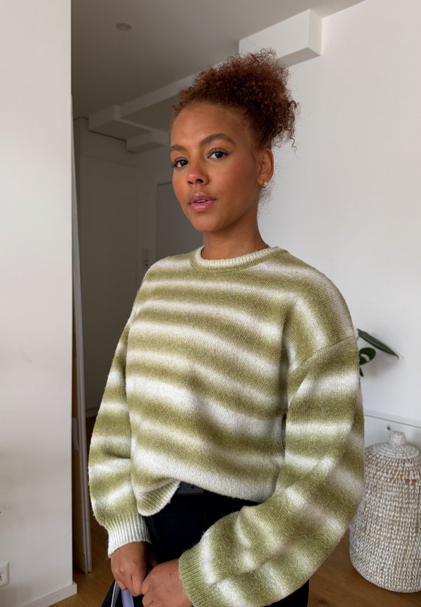 Femme aux cheveux bouclés en chignon haut, portant un pull rayé vert et blanc, debout à l’intérieur près d’un mur blanc et d’un panier tressé.