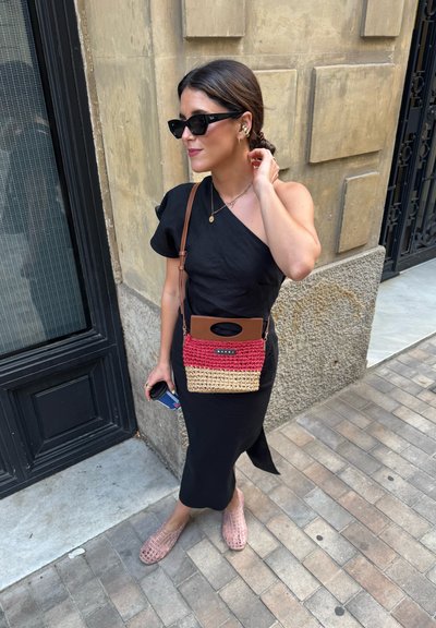 Mujer con vestido negro de un solo hombro, gafas de sol y bolso de mimbre, de pie en una acera de la ciudad sosteniendo una pequeña taza de café.