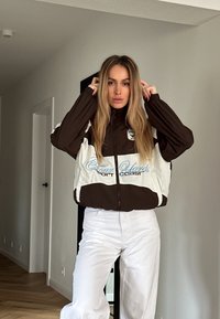 Brun og hvid zip-up jakke med elastiske manchetter, der har et logo og tekstdetalje. Parret med hvide bukser, sat mod en neutral baggrund.