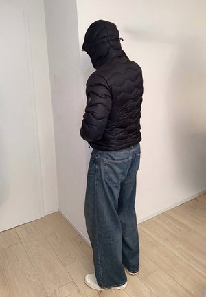 Persona de espaldas en una esquina, vistiendo una chaqueta acolchada con capucha negra, vaqueros anchos y zapatillas blancas sobre suelo de madera clara en el interior.