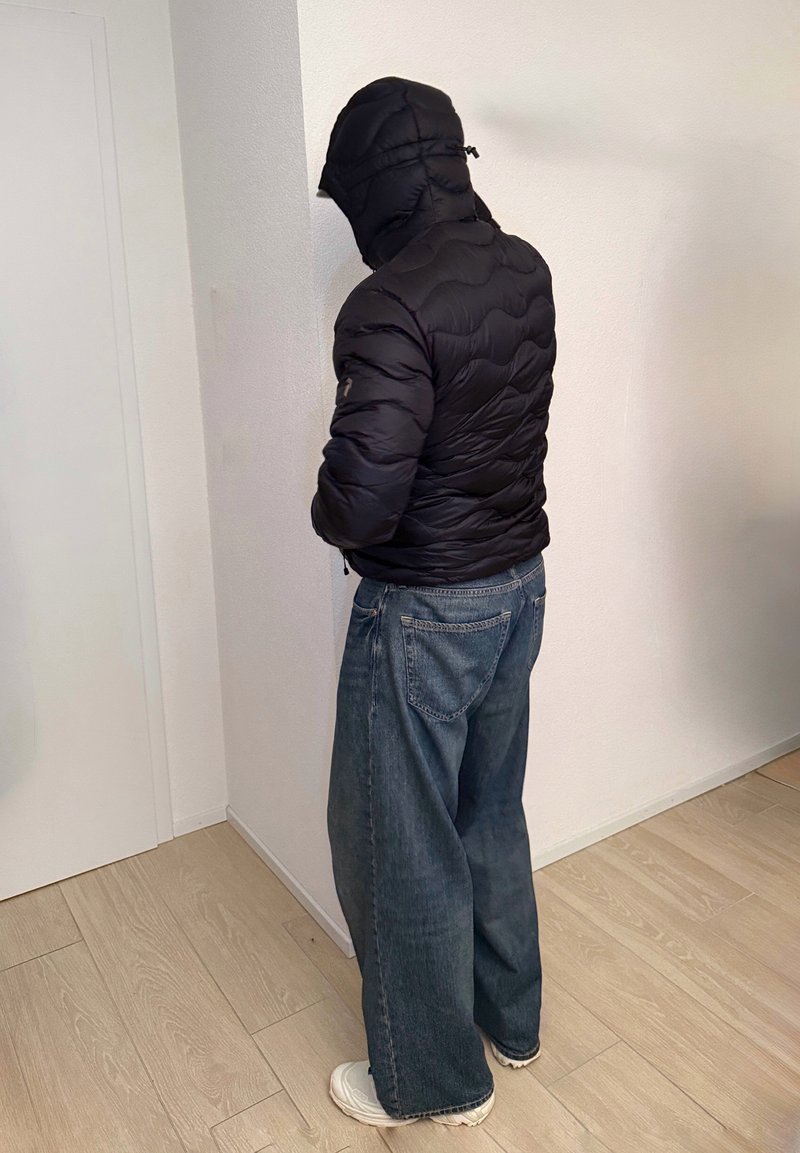 Persoon die naar een hoek toe staat, gekleed in een zwarte gewatteerde jas met capuchon, brede spijkerbroek en witte sneakers op een lichte houten vloer binnen.