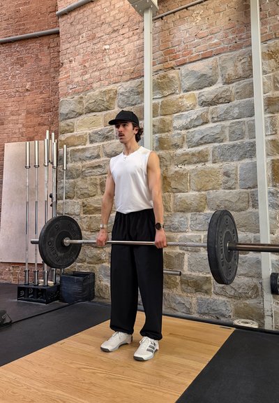 Hombre con gorra negra, camiseta blanca sin mangas y pantalones negros levantando una barra en un gimnasio con paredes de ladrillo y piedra.