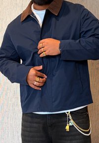 Veste bleu marine avec un col en velours côtelé marron, tissu résistant à l'eau, coupe droite et fermeture éclair devant. Accessoires comprenant plusieurs bagues et chaînes.