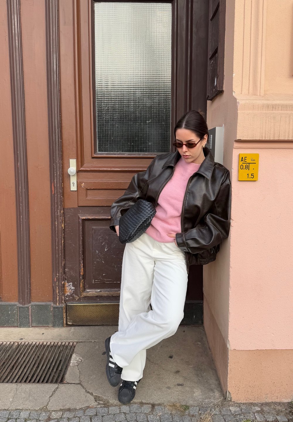 Brown leather jacket over a pink sweater, white wide-leg trousers, and black sneakers. Carrying a woven black bag. Standing by a door.