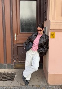 Brown leather jacket over a pink sweater, white wide-leg trousers, and black sneakers. Carrying a woven black bag. Standing by a door.