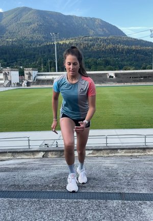 La camiseta atlética presenta un degradado de azul, gris y coral, combinada con pantalones cortos negros y zapatillas blancas. El corredor está ascendiendo por una pista.