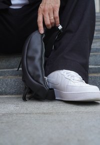 Melnas ādas jostas soma ar teksturētu apdari, sānu rāvējslēdzis un regulējama siksna, novietota blakus baltam sportam uz pelēkiem soļiem.
