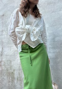 Femme portant un chemisier à fleurs blanches noué à l'avant, un top blanc en dessous, et une jupe verte, se tenant devant un mur texturé.