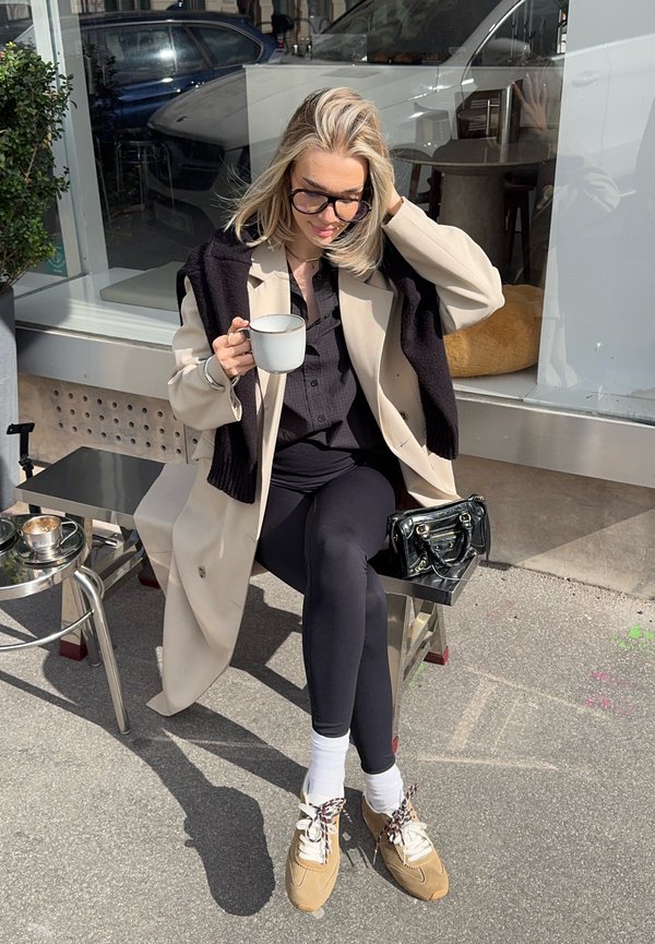 Lyshåret kvinde med briller, beige frakke og sorte leggings sidder udendørs på en metalbænk og holder en hvid krus med en sort pung ved siden af sig.