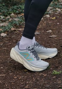 Tênis atléticos cinzas com detalhes em teal, parte superior texturizada, colarinho acolchoado e solas brancas grossas. Usados com meias cinzas e leggings pretos.