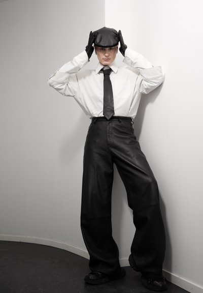 Persona con camisa blanca, corbata negra, pantalones anchos de cuero negro, guantes y gorra negra, de pie contra una pared blanca curvada.
