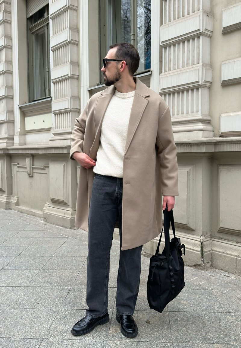 Manteau en laine beige, pull texturé blanc, jean gris, chaussures en cuir noires et sac en nylon noir. L'arrière-plan montre des détails architecturaux.