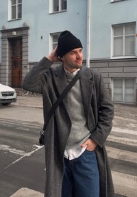 Szürke halszálkamintás kabát, világosszürke pulóver, fehér ing, kék farmernadrág és fekete sapka. A személy egy utcán áll egy világoskék épület előtt.