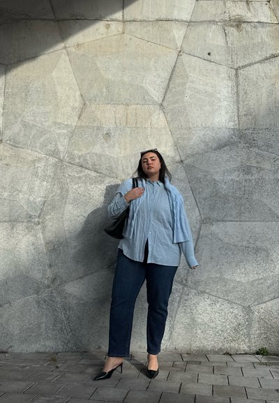Camisa de botón azul claro combinada con un cárdigan de punto gris, jeans oscuros y tacones negros frente a una pared de piedra gris texturizada. Bolso negro.
