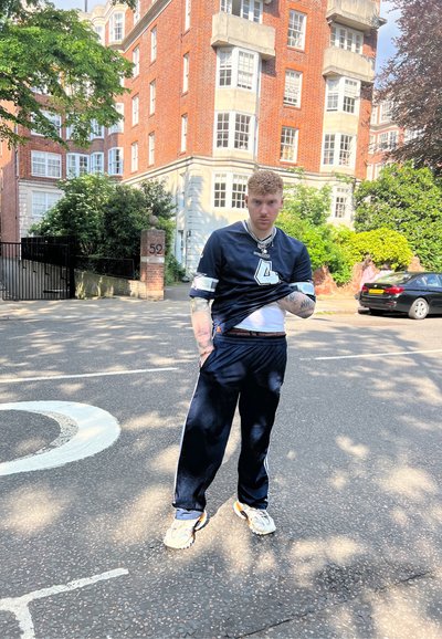 Chándal azul marino con franjas blancas; camiseta negra de manga corta con el número 4; zapatillas gruesas; entorno urbano con edificios de ladrillo.