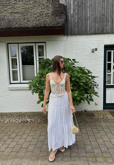 Una mujer que lleva una blusa de encaje blanca y una falda larga, con gafas de sol y un bolso en forma de corazón, se encuentra en un camino de adoquines cerca de una casa de ladrillo blanco.