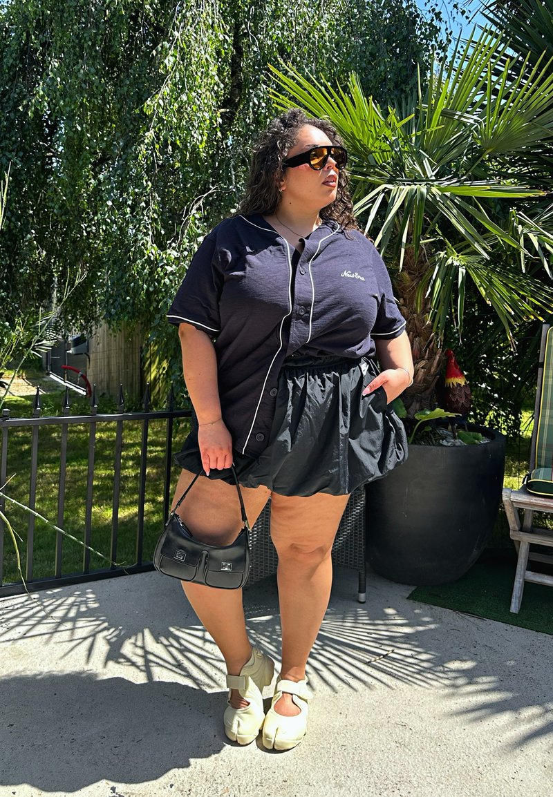 Femme portant une chemise noire, une jupe noire, des chaussures blanches à orteils séparés et des lunettes de soleil se tient dehors en tenant un petit sac noir près des plantes et d'une clôture.