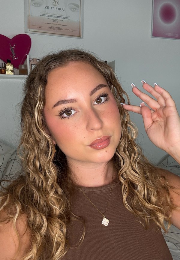 Jeune femme aux cheveux blonds bouclés portant un haut marron sans manches et un collier pendentif, touchant ses cheveux d'une main à l'intérieur.