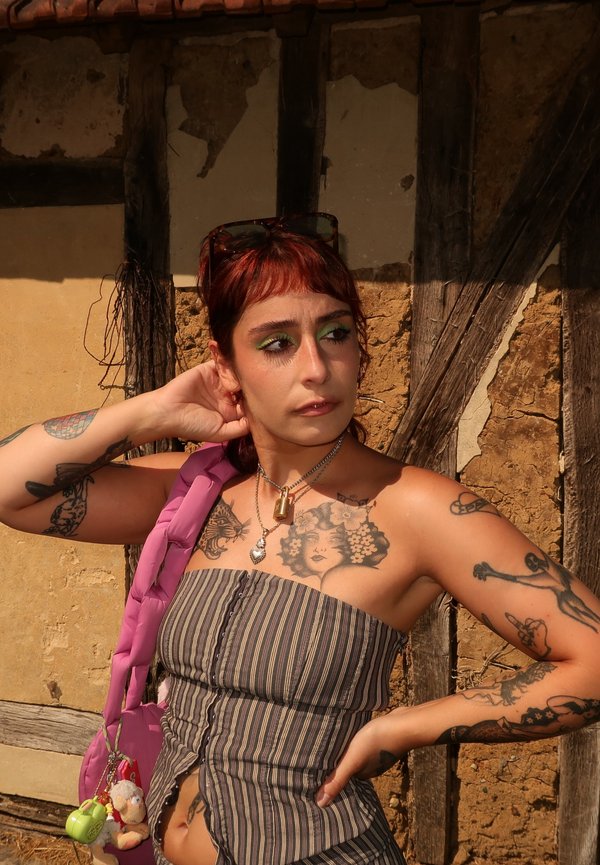 Joven mujer con cabello rojo y tatuajes, vestida con un top a rayas sin tirantes, de pie contra una pared desgastada de madera y yeso bajo la luz del sol.