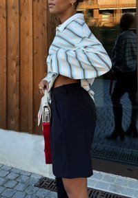 Stripete langermet skjorte i beige, blått og brunt, kombinert med svarte skreddersydde shorts. En lys veske med rødt stropp holdes i hånden.