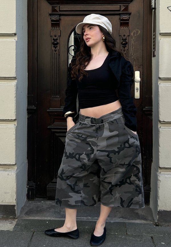 Vrouw in een witte hoed, zwart cropped topje, zwart jasje, grijze camouflage wijde broek en zwarte platte schoenen staand voor een donkere houten deur.