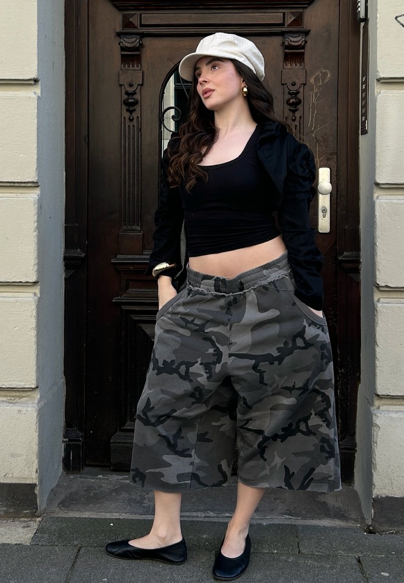 Mujer con sombrero blanco, top negro corto, chaqueta negra, pantalones anchos de camuflaje gris y zapatos planos negros, de pie frente a una puerta de madera oscura.