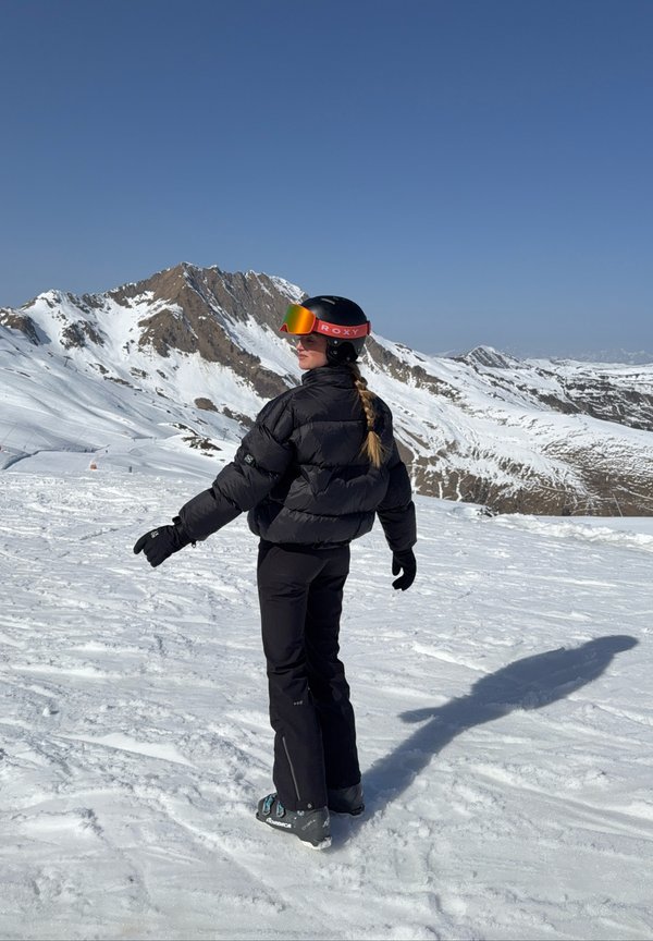 Vrouw in zwarte skikleding en helm staat op sneeuw en kijkt over haar schouder met besneeuwde bergen en een heldere blauwe lucht op de achtergrond.