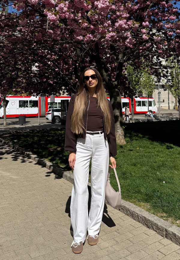 Mujer con gafas de sol, top oscuro, pantalones blancos sosteniendo un bolso beige, de pie bajo un árbol con flores rosas cerca de un tranvía rojo y blanco.