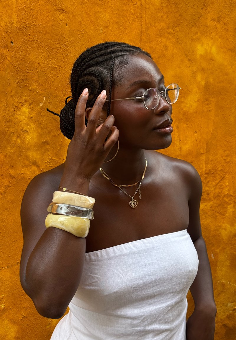 Lunettes transparentes, un haut blanc bustier et plusieurs bracelets joncs en crème et argent. L'arrière-plan est un mur jaune vif.