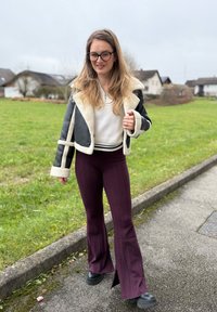 Zwart en crèmekleurig shearlingjack, witte geribbelde trui en wijnrode, hooggetailleerde uitlopende broek met kabelgebreide textuur. Zwarte enkellaarsjes.