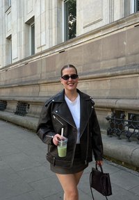 Jachetă de piele neagră pentru bicicliști cu fermoare argintii, tricou alb, fustă mini neagră. Ține o băutură și poartă o geantă neagră împletită.