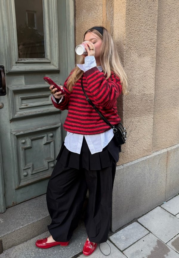 Tânără în pulover cu dungi roșii și pantaloni negri bea dintr-o ceașcă în timp ce ține un telefon, stând lângă un perete texturat și o ușă verde.